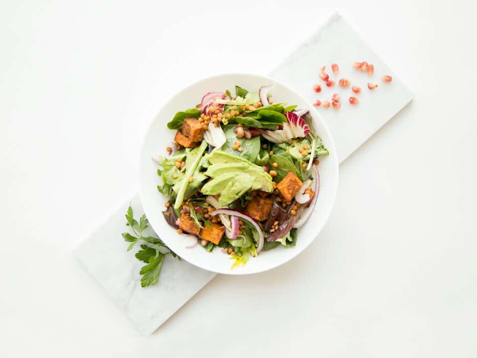 Bol cu salată verde, avocado, linte, cartofi dulci copți și ceapă roșie, pe o masă de marmură
