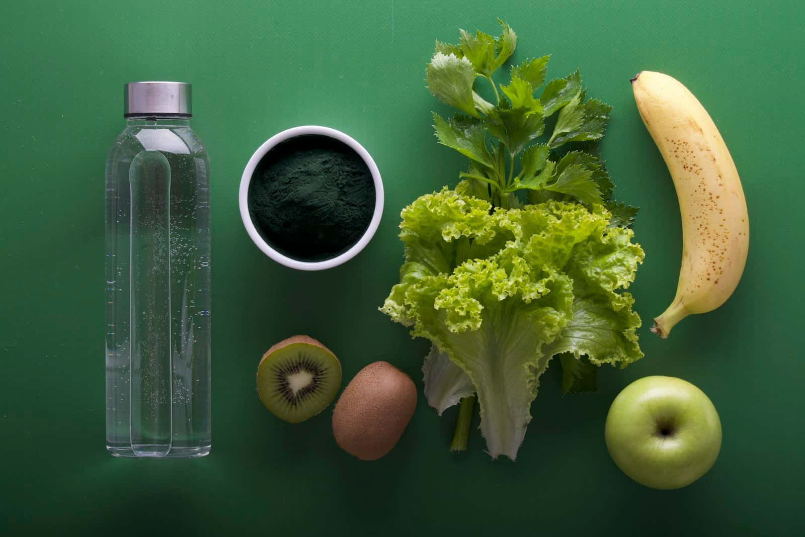 Sticlă de apă, kiwi, salată verde, țelină, o banană și un măr verde așezate pe un fundal verde